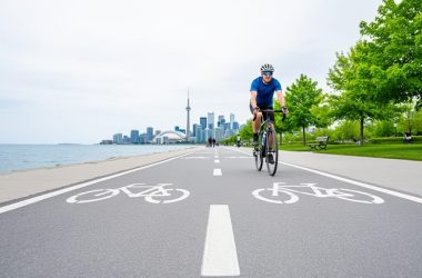 A cyclist rides along Toronto’s waterfront multi-use trail with trees and the distant downtown skyline in the background.